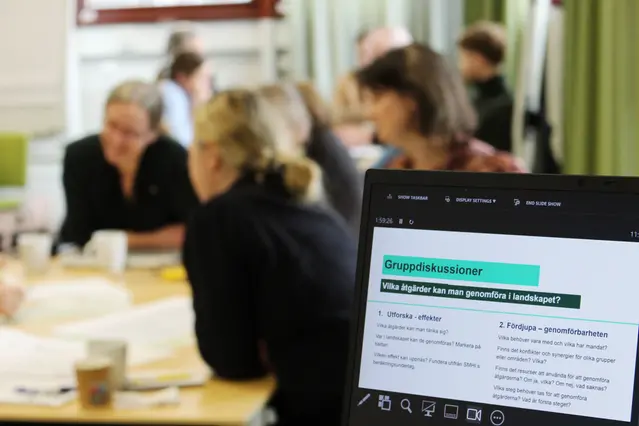 I framgrunden syns i datorskärm med diskussionsfrågor, framför en grupp personer som diskuterar vid ett bord.