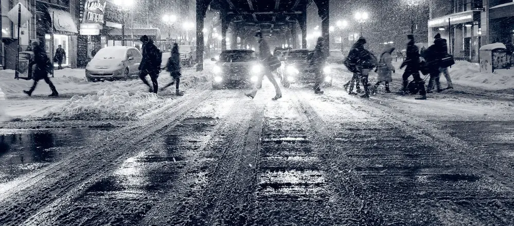 Svartvit bild på människor som korsar en väg i snöfall med bilar i bakgrunden