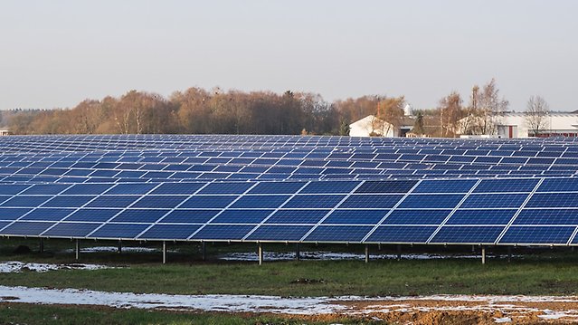 Solpaneler i långa rader på öppen mark, i bakgrunden industribyggnader. 